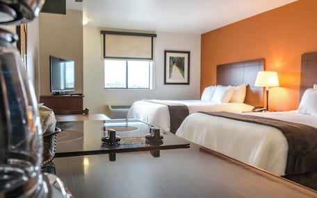 A hotel room with two beds, a TV, a window with blinds, a framed picture on the wall, and a bedside lamp.
