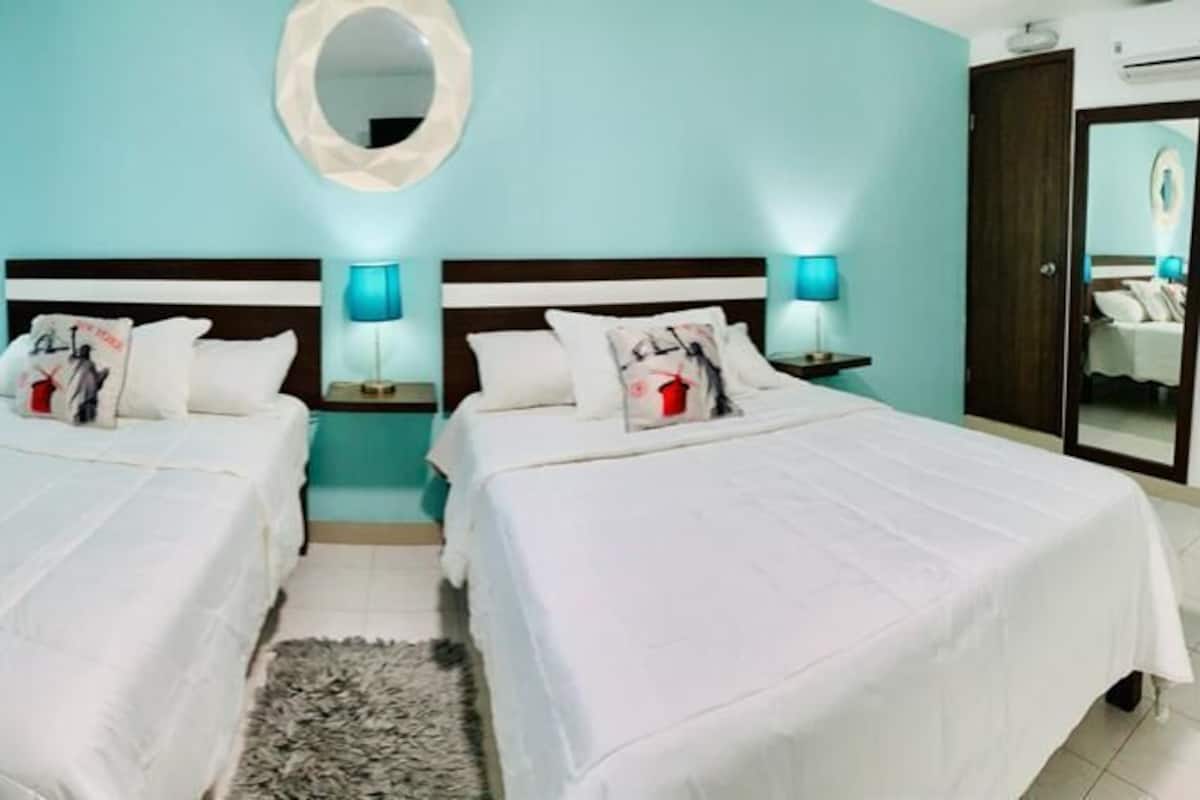 A hotel room with two beds, a round mirror, a small rug, and a nightstand with a lamp.
