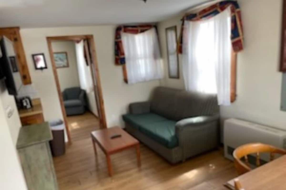 Et kompakt opholdsrum med en sofa, et lille bord og en stol. Der er en døråbning til et andet rum, og en radiator er synlig på væggen.