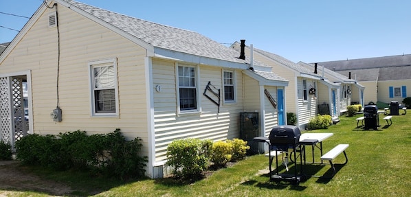 A row of single-story, yellow houses with white trim, each featuring a small front yard with a picnic table and grill.