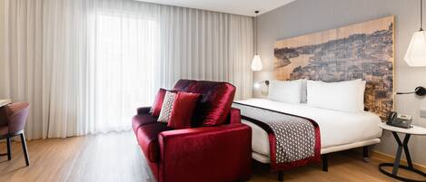 A hotel room with a bed, a red sofa, a desk, and a cityscape painting on the wall.