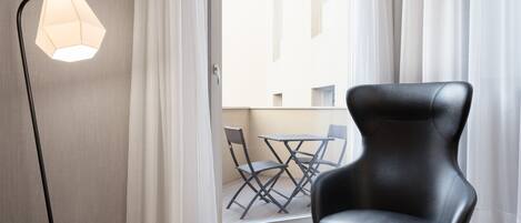 A modern room with a black leather chair, a floor lamp, and a view of an outdoor balcony with a table and chairs.
