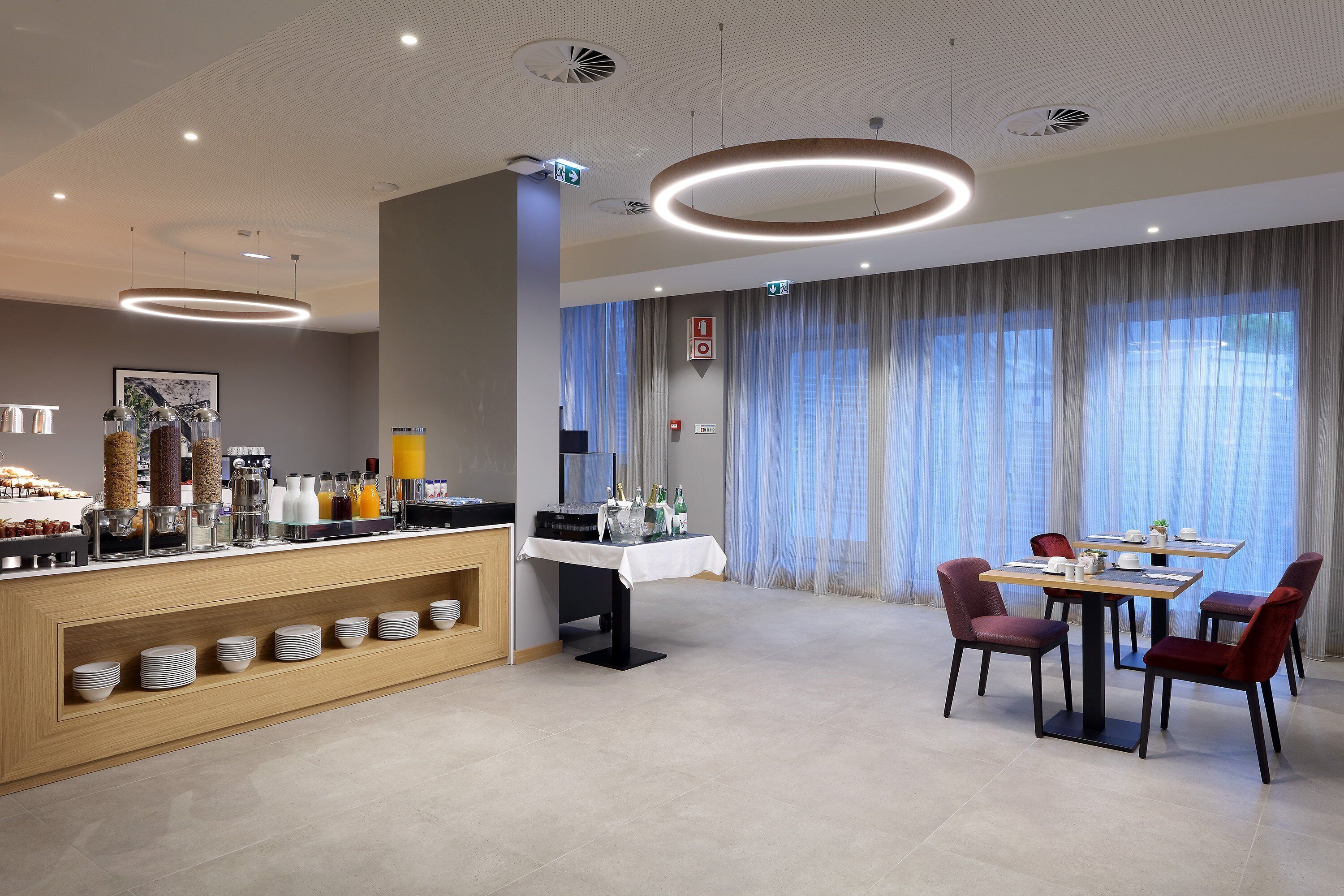A modern hotel dining area with a buffet, a table with chairs, and a large window with curtains.