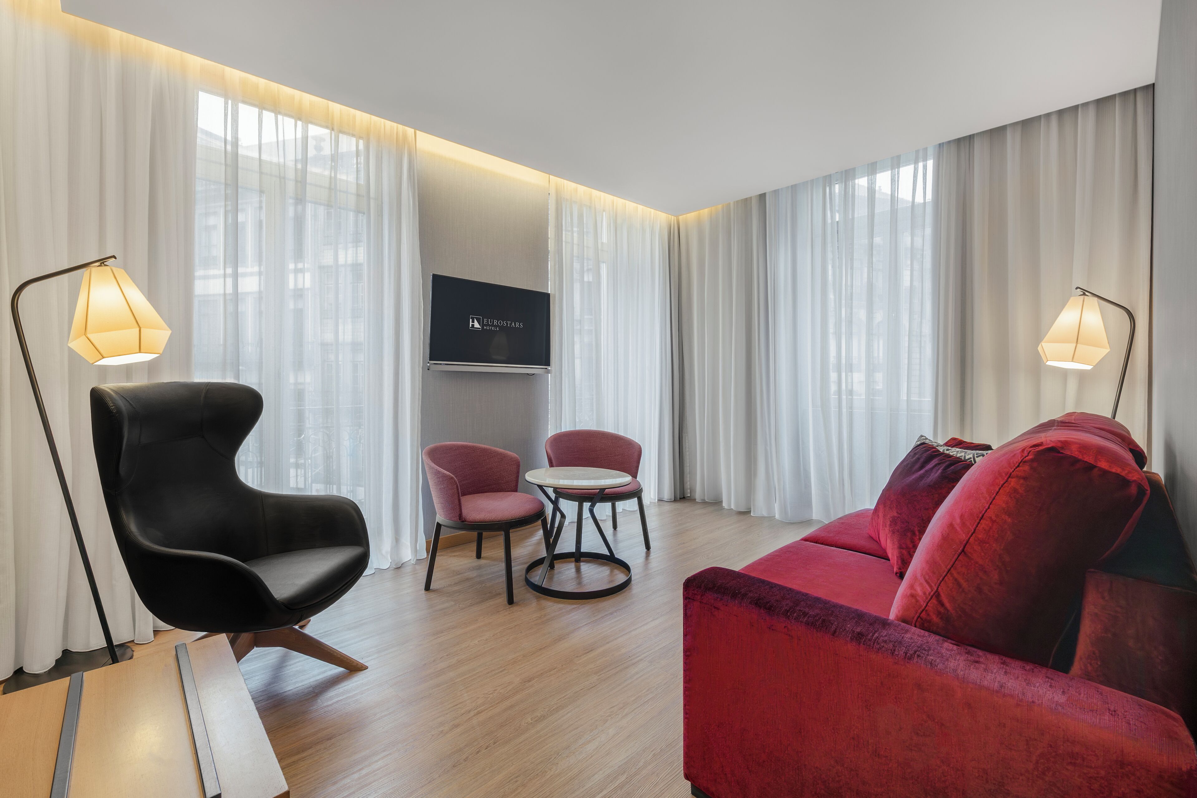 Une chambre d’hôtel moderne avec un canapé rouge, un fauteuil noir et deux chaises roses disposées autour d’une petite table. Un téléviseur à écran plat est fixé au mur.