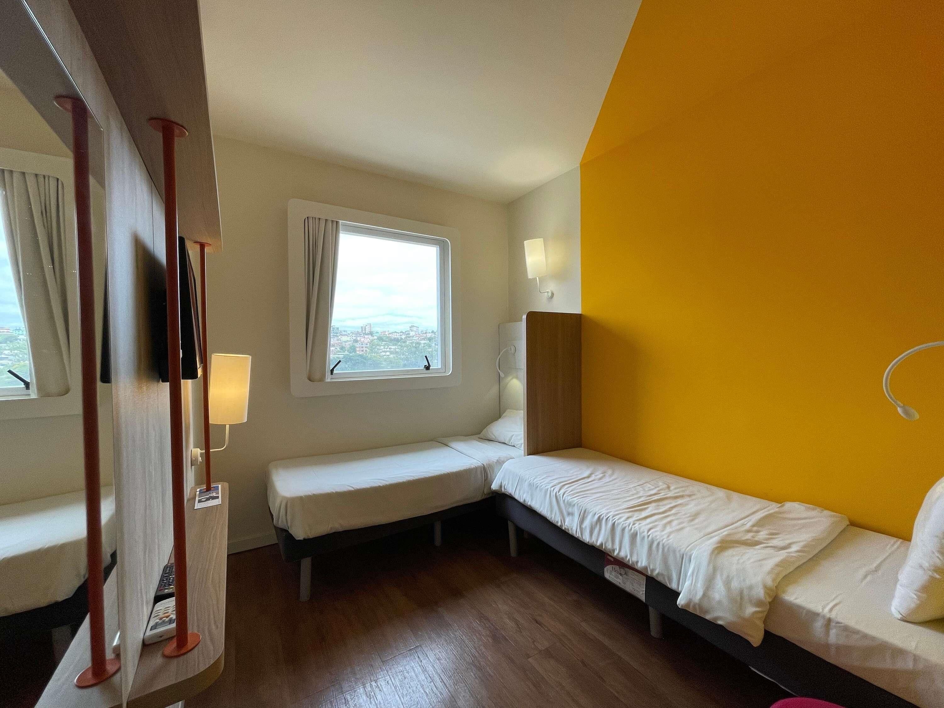Una habitación de hotel pequeña con dos camas individuales, una ventana con cortinas, una mesita de noche con lámpara y vistas a la ciudad.