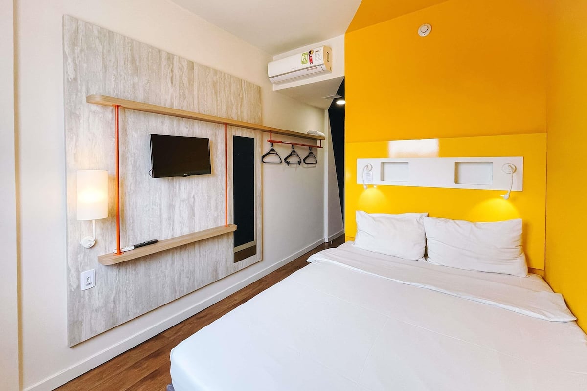 Quarto de hotel com cama, televisão montada, prateleira com abajur e guarda-roupa com roupas penduradas.