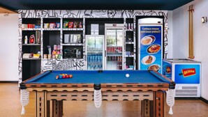 Ein Billardtisch mit blauer Spielvliesdecke, umgeben von Regalen voller Snacks und Getränke, sowie ein Verkaufsautomat mit Lebensmitteln.