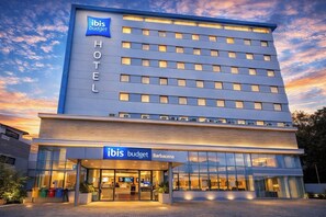 A modern hotel with a prominent 'ibis budget' sign, a glass facade, and a 'Barbacena' label.