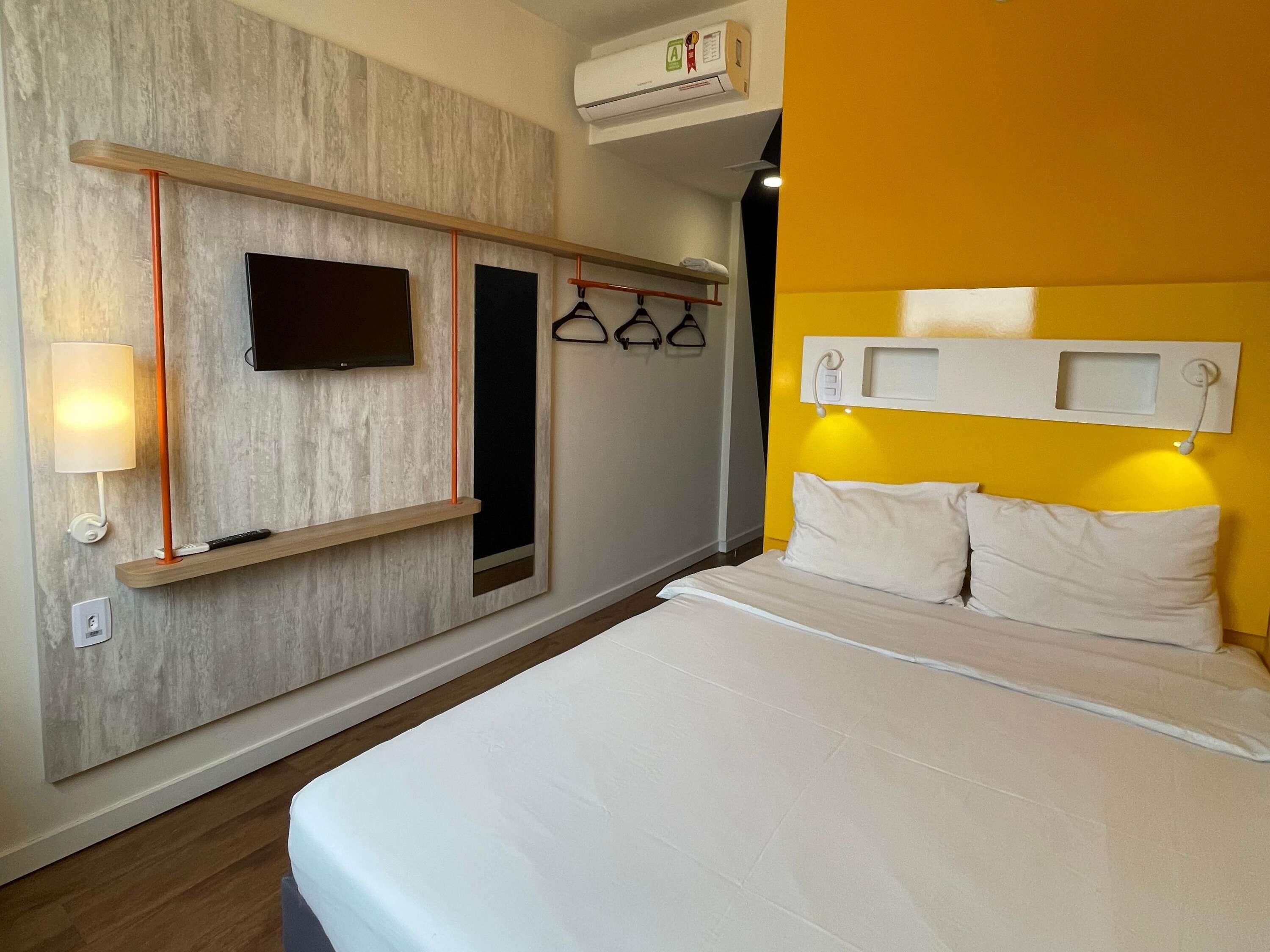 Una habitación de hotel con cama, televisor montado, calentador en la pared y perchero.