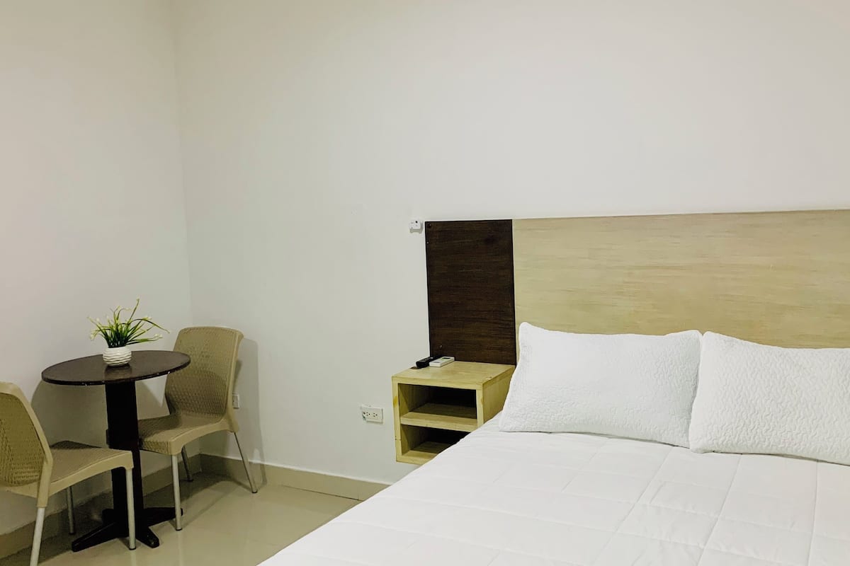 Ein Hotelzimmer mit einem Bett, einem kleinen runden Tisch mit Topfpflanze, zwei Stühlen und einem Nachttisch mit Fernbedienung.