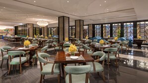Uma ampla sala de jantar com várias mesas e cadeiras, com um candelabro e janelas grandes com painéis de vidro decorativos.