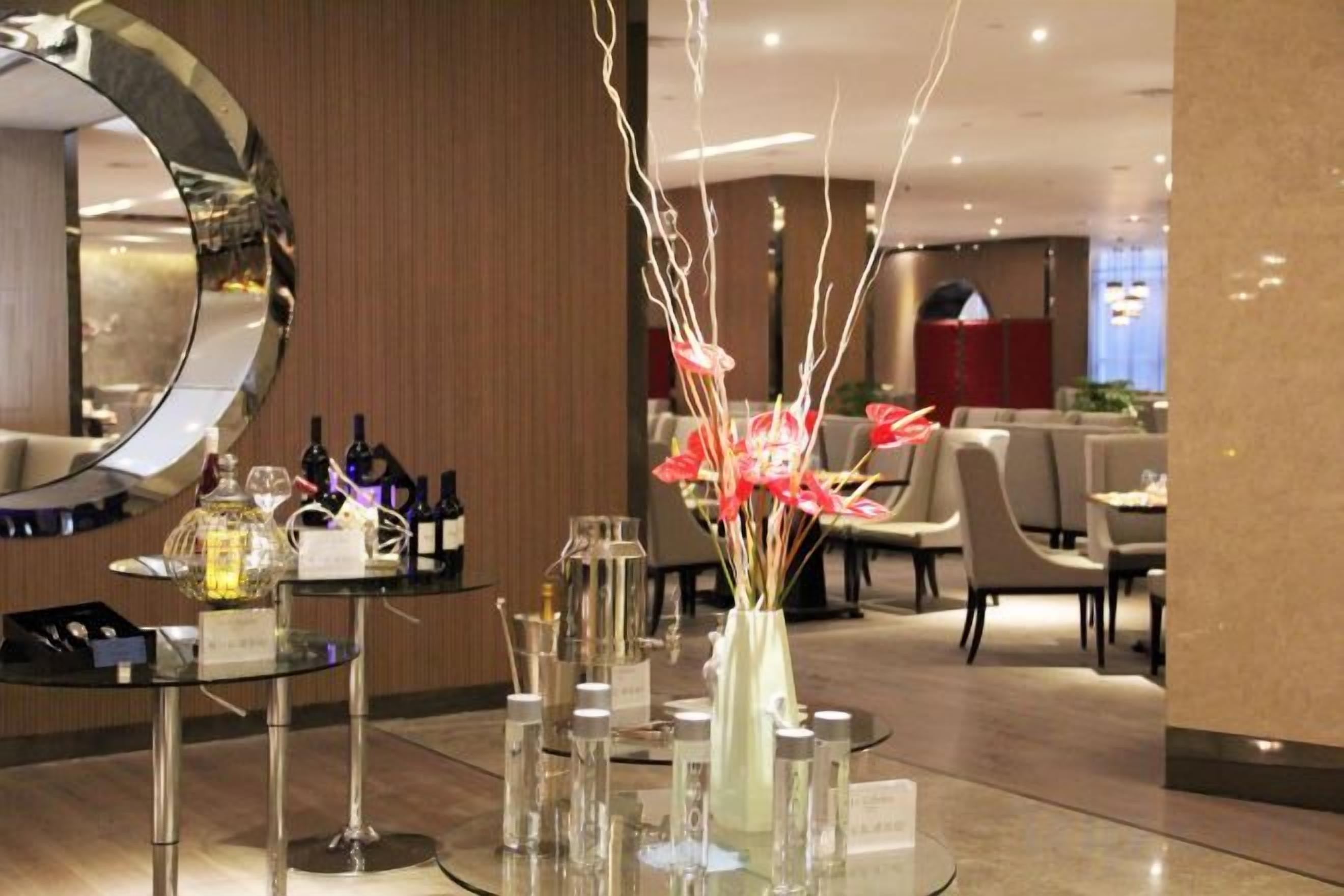 A hotel lobby with a round mirror, a glass table with a vase of flowers, and a bar area with bottles.
