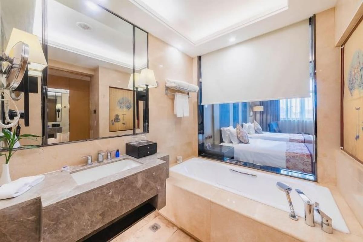 A modern bathroom with a large mirror, dual sinks, a bathtub, and a view of a bedroom with a bed and curtains.