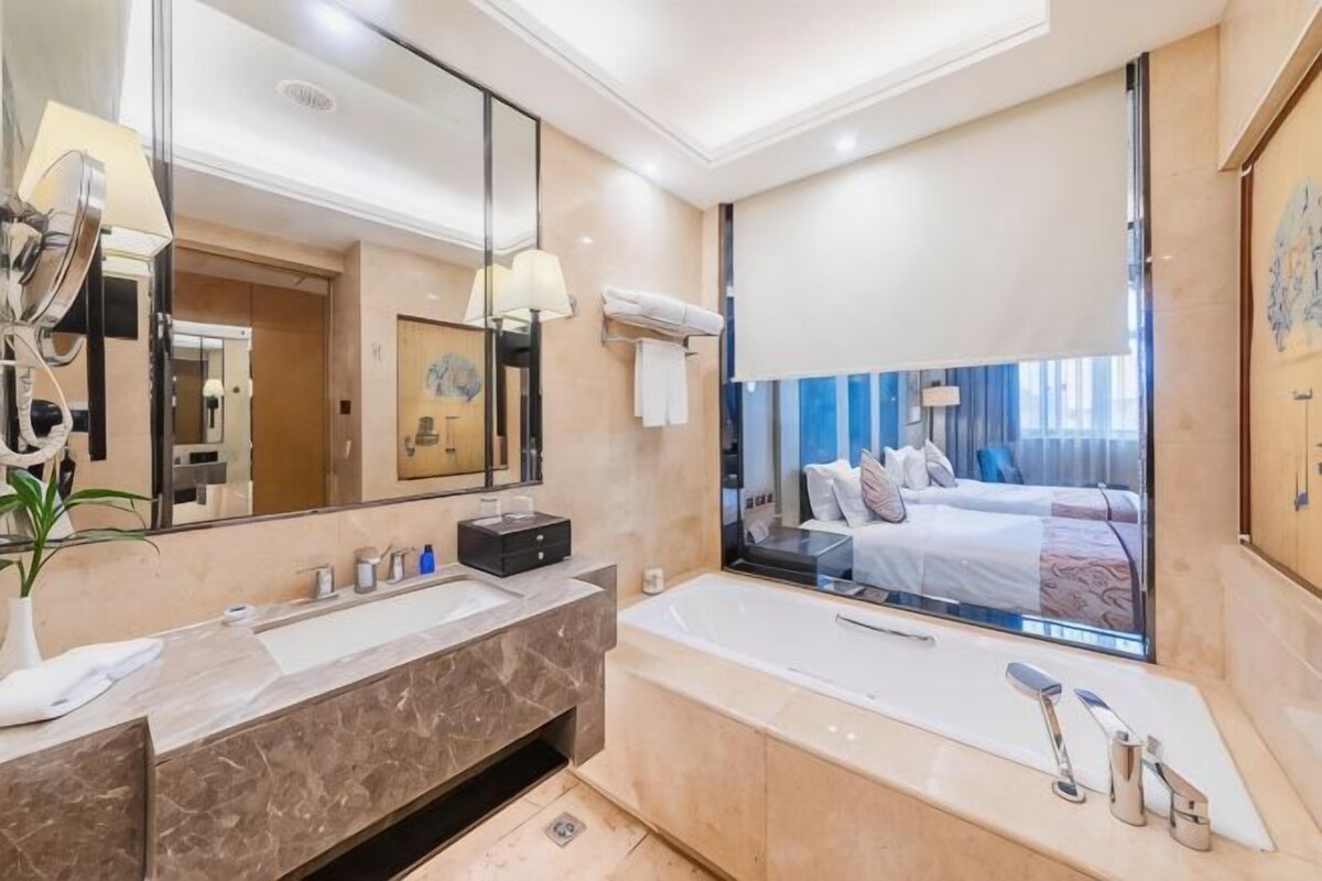 Ein modernes Badezimmer mit einem großen Spiegel, einem Waschtisch mit zwei Becken, einer Badewanne und einem großen Fenster mit Blick auf ein Schlafzimmer.
