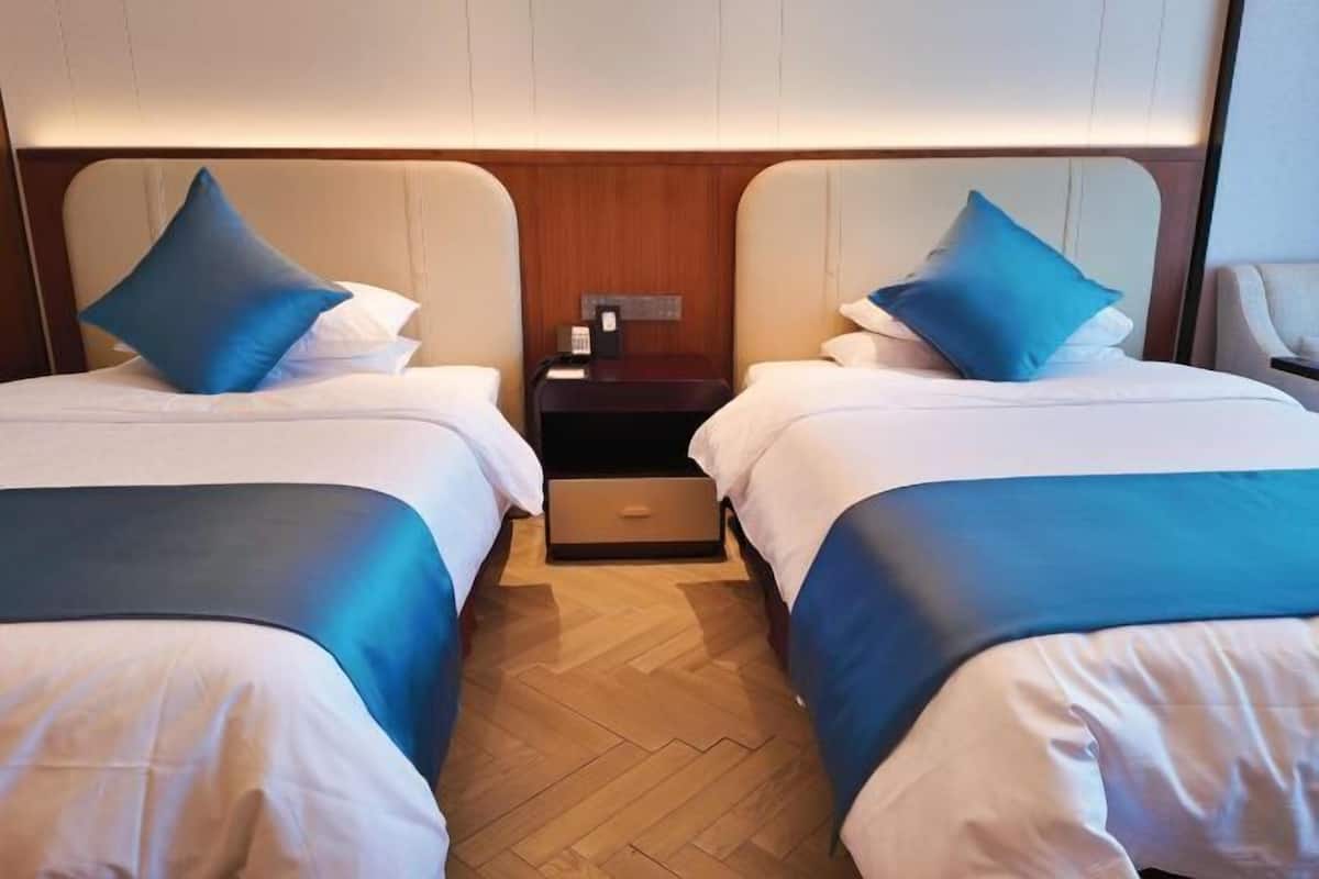 Ein Hotelzimmer mit zwei Betten, jedes mit einem blauen Kopfkissen und weißer Bettwäsche. Es gibt ein Holz-Kopfteil, einen Nachttisch mit Telefon und eine Schublade.