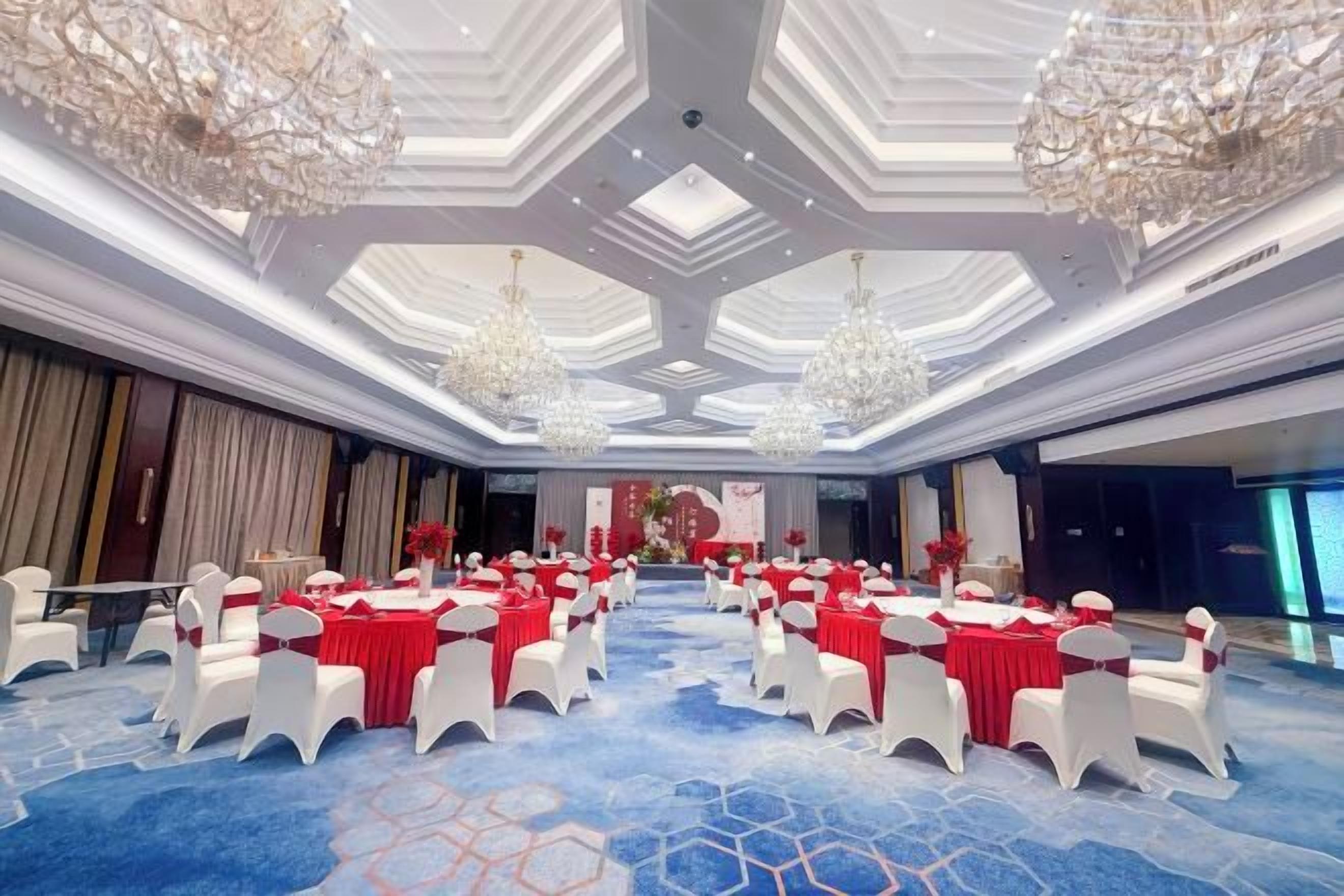 A large conference room with round tables and white chairs, chandeliers, and a blue carpet.