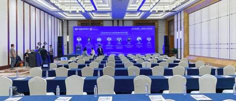 A conference room with rows of tables and chairs, a stage with a presentation screen, and a panel of speakers.