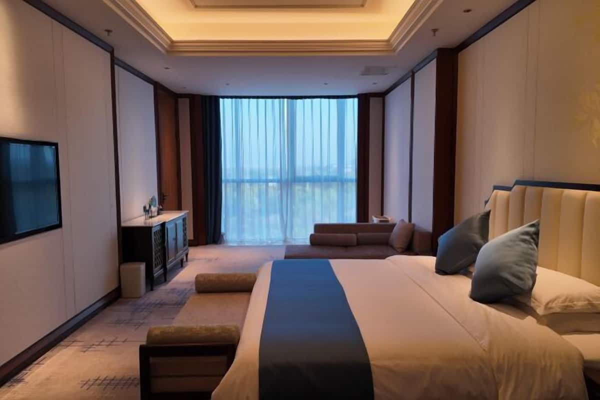 Ein Hotelzimmer mit einem großen Bett, einem Fernseher, einem Schreibtisch und Blick auf die Stadt durch transparente Vorhänge.