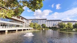 Ein modernes Gebäude mit Glasfassade, einer Brücke über ruhigem Wasser und umgebender Bepflanzung.