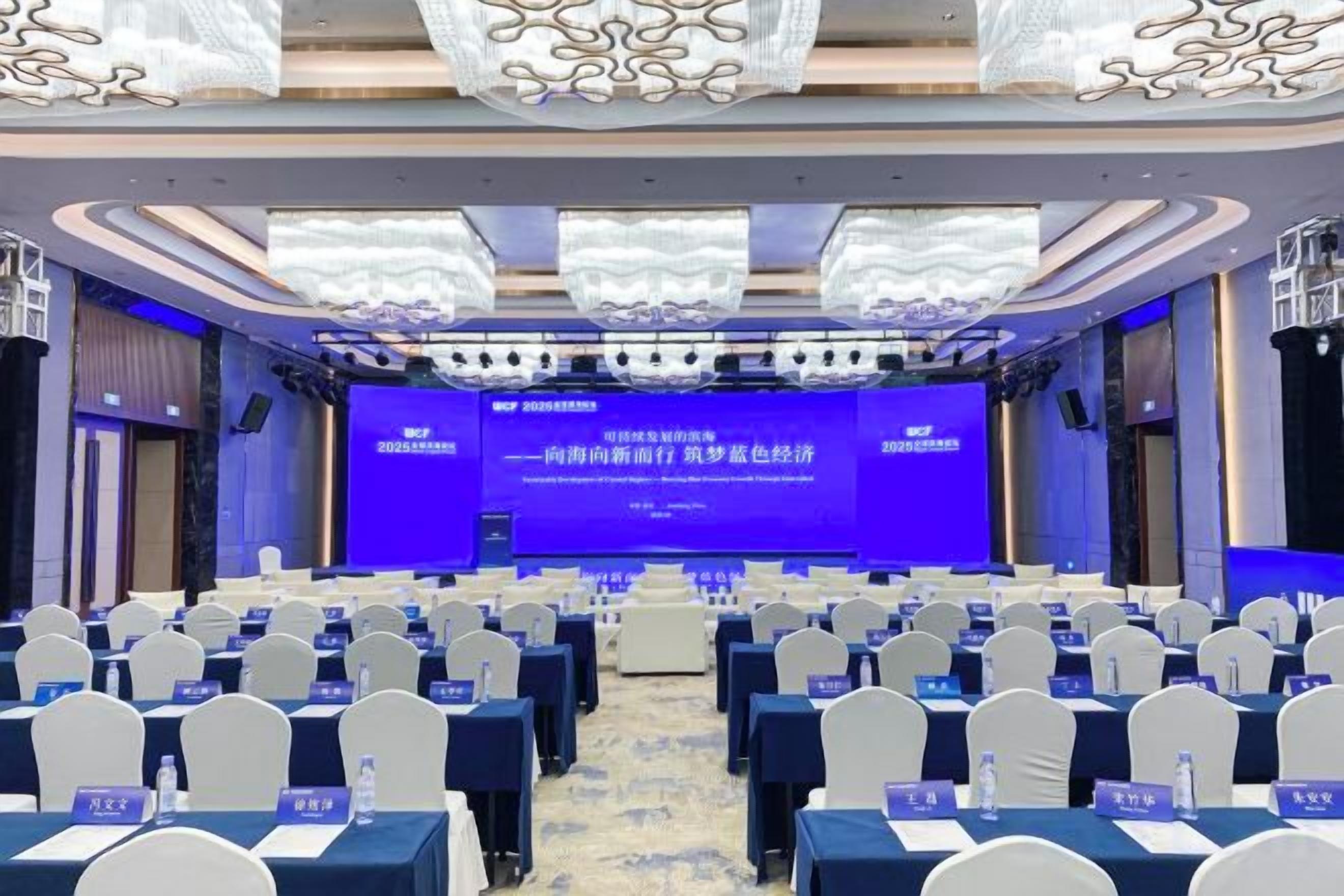 Un salón de conferencias con filas de mesas y sillas, una pantalla grande al frente y una alfombra azul.