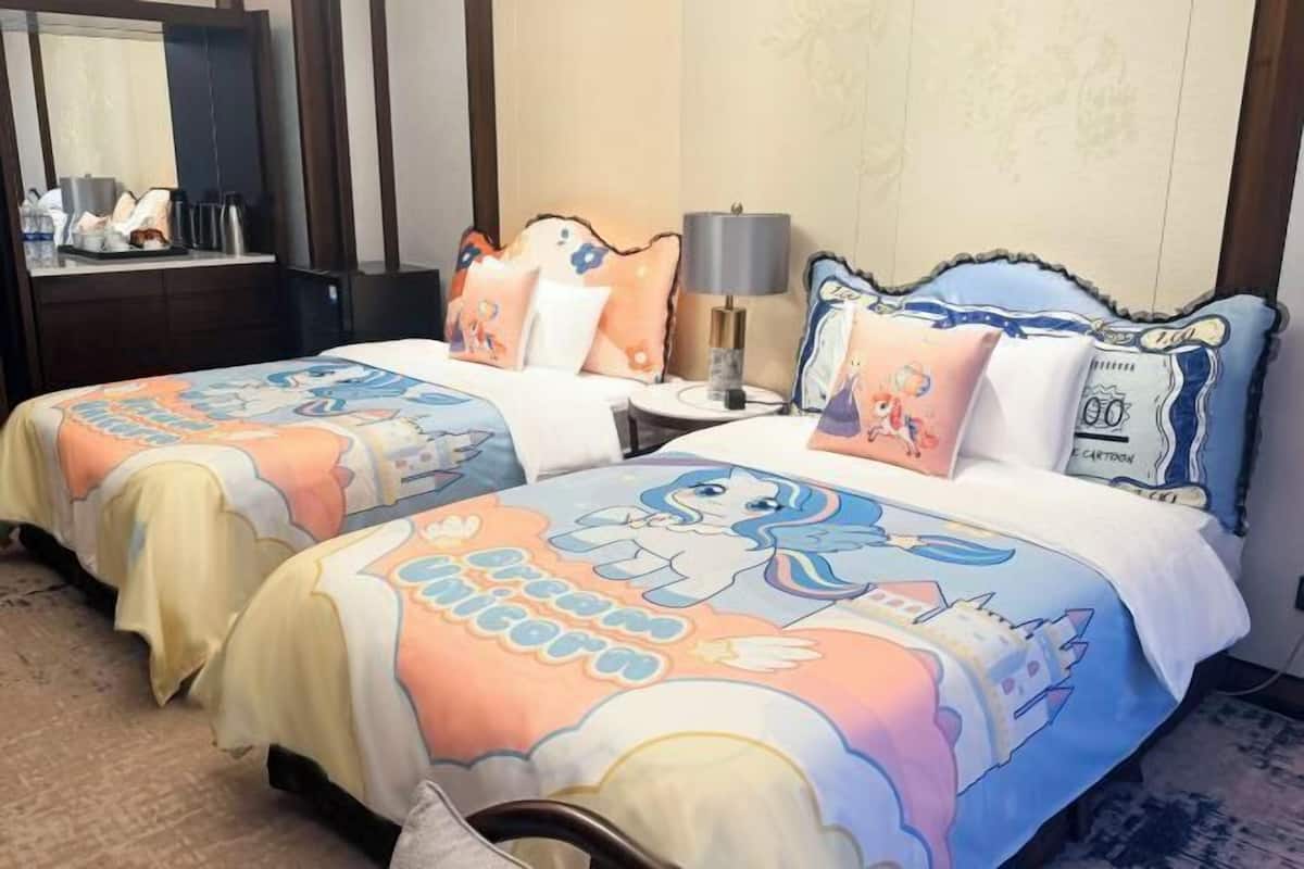 Ein Hotelzimmer mit zwei Betten, jedes mit einer bunten Bettdecke im Einhorn-Design, einem Nachttisch mit Lampe und einem Spiegel an der Wand.