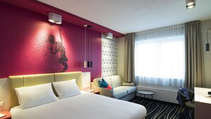 Une chambre d’hôtel avec un lit, un canapé, une petite table ronde, un bureau et une grande fenêtre avec des rideaux.