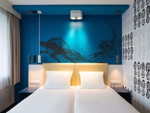 A hotel room with a large bed, two bedside tables, a wall mural, and a ceiling light fixture. - ibis Styles Haarlem City (Haarlem)