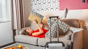 Une personne est allongée sur un canapé gris, une valise à ses côtés, et lit un livre dans une pièce dont le mur est décoré d’un motif et qui possède une fenêtre avec des rideaux.