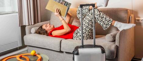 A person lying on a grey sofa with a suitcase, reading a book, in a room with a patterned wall and a window with curtains.