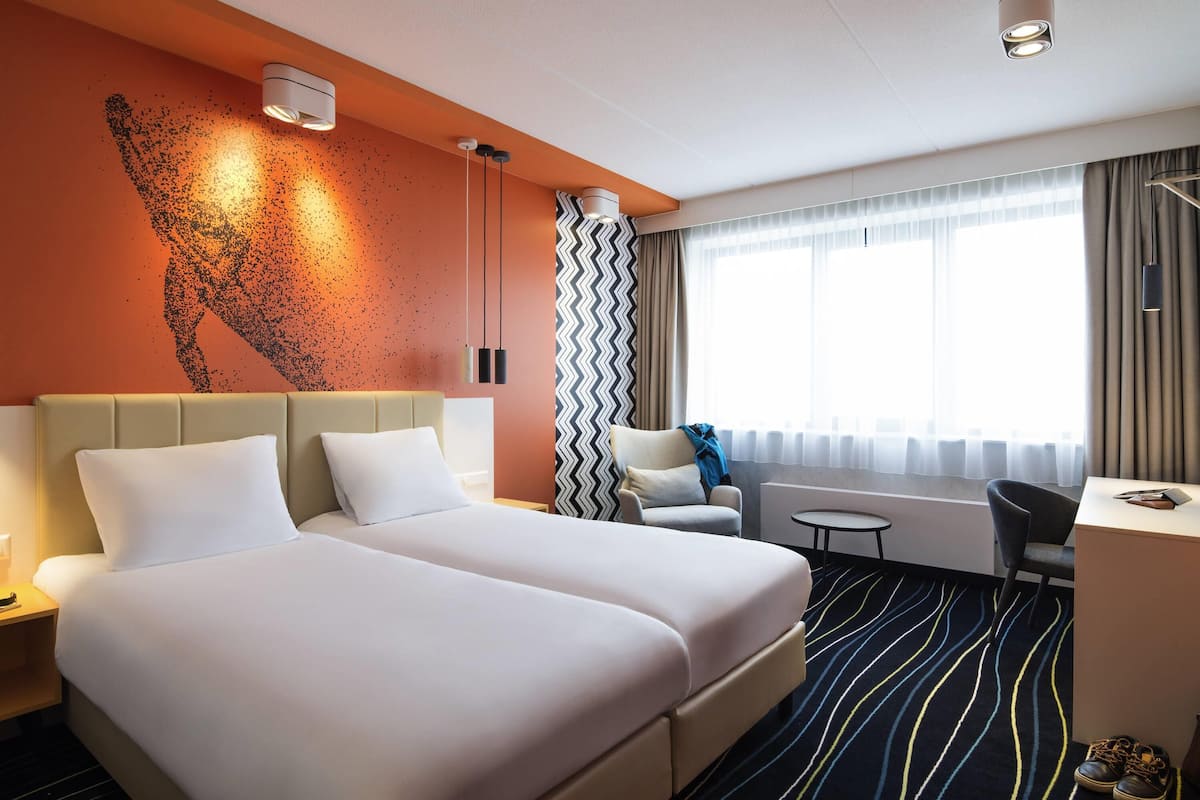 A hotel room with two beds, a desk, a chair, and a window with curtains.