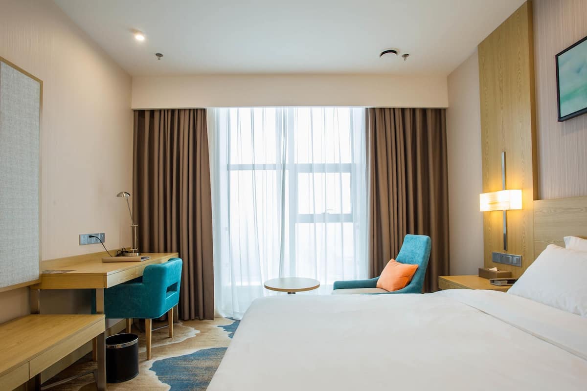 A hotel room with a large bed, a desk, a chair, a small table, and a nightstand with a lamp.
