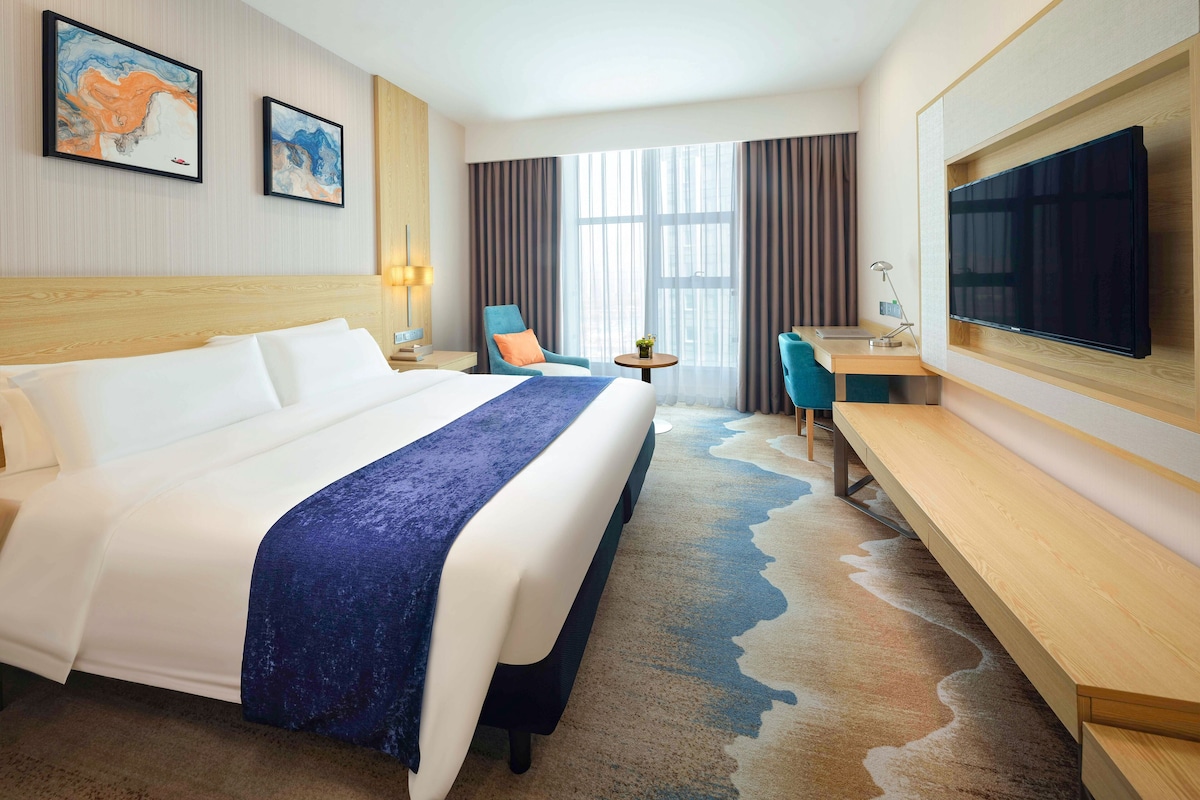 A hotel room with a large bed, a desk, a chair, a TV, and artwork on the walls.