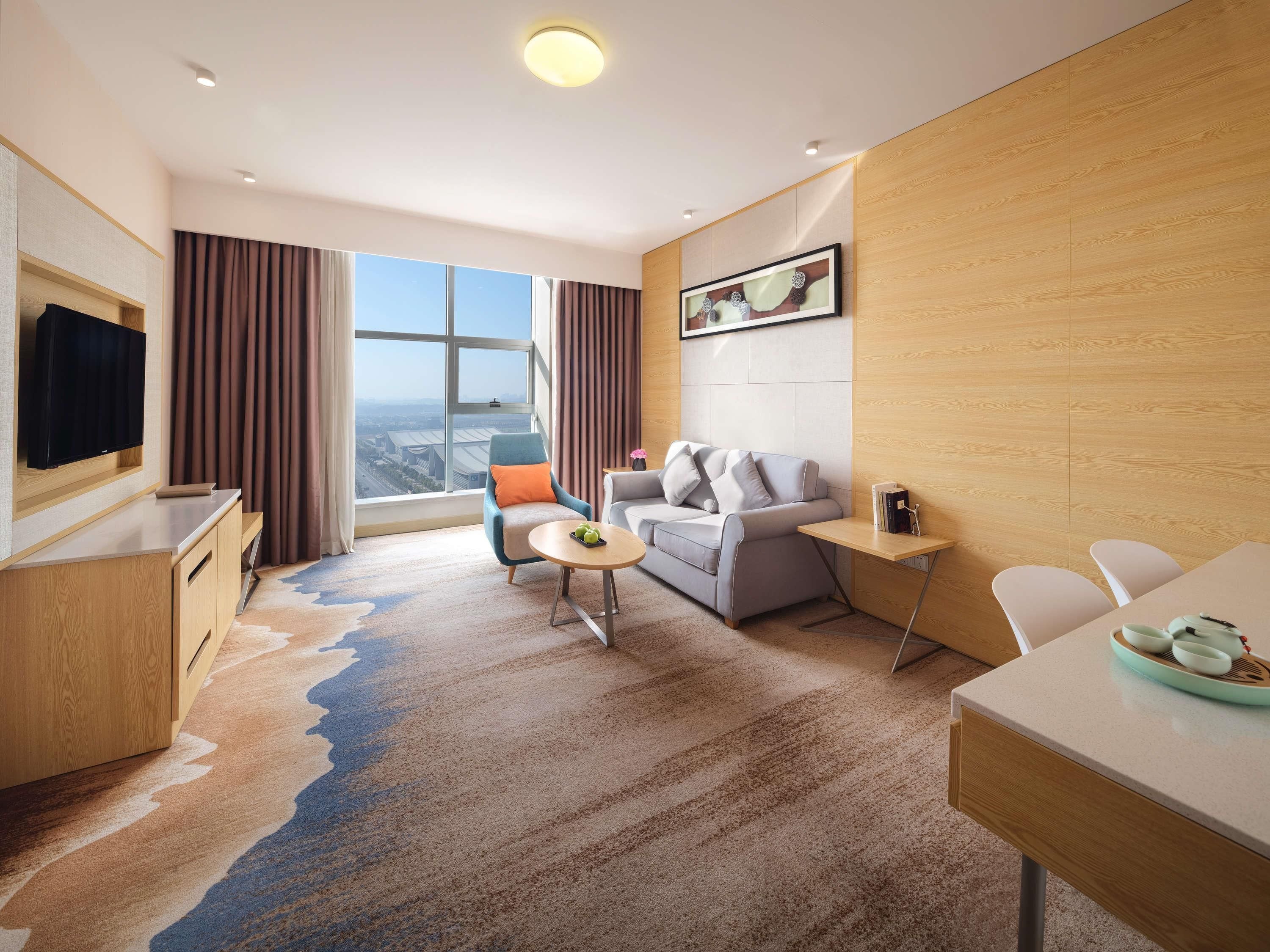 Ein modernes Hotelzimmer mit großem Fenster, einer Couch, einem Esstisch und einem Fernseher.