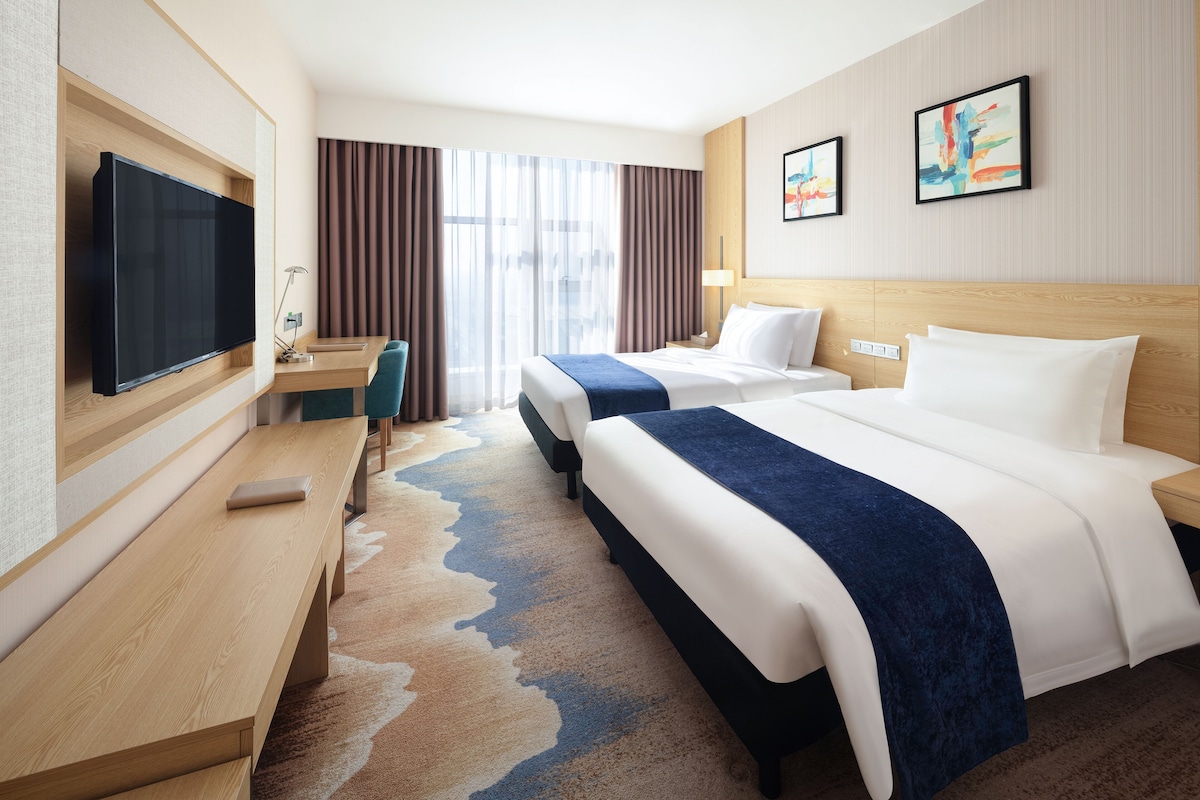 A hotel room with a flat-screen TV, a desk with a chair, two beds, a large window with curtains, and two framed artworks on the wall.