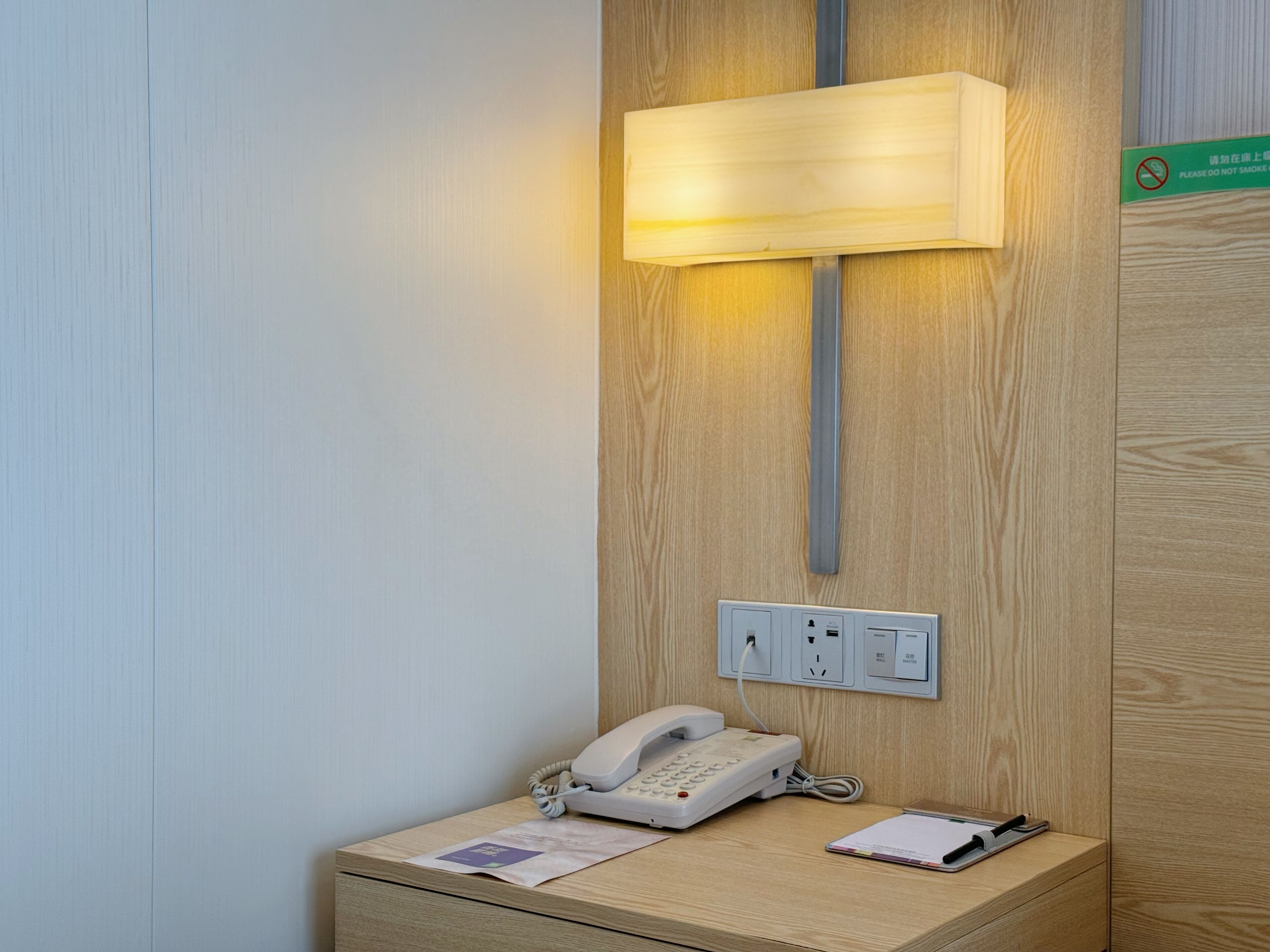 Habitación de hotel con teléfono en la mesita de noche, lámpara y toma de corriente en la pared.