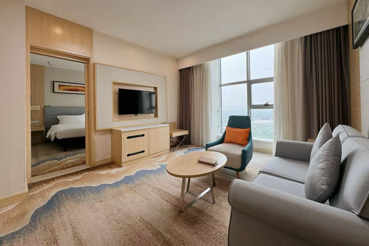 A hotel room with a bed, a TV mounted on a wooden cabinet, a seating area with a sofa and armchair, and a large window offering a city view.