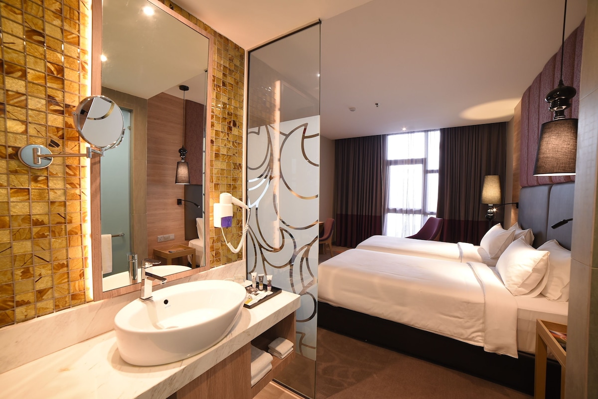 A modern hotel room with a large bathroom vanity, a large mirror, a bed with white linens, and a view of the city through the window.