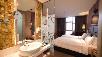 A modern hotel room with a large bathroom vanity, a large mirror, a bed with white linens, and a view of the city through the window.
