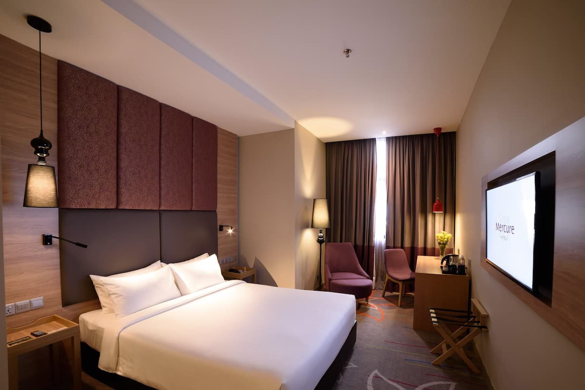 A hotel room with a bed, a desk, a chair, a TV, and a wall-mounted screen displaying the hotel name.