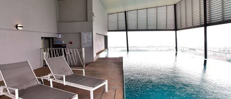 An indoor swimming pool with clear blue water, surrounded by white lounge chairs and a high ceiling with large windows.