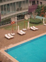 Piscina con sillas de descanso y sombrillas, rodeada por un suelo de ladrillo y paredes de cristal de un edificio.