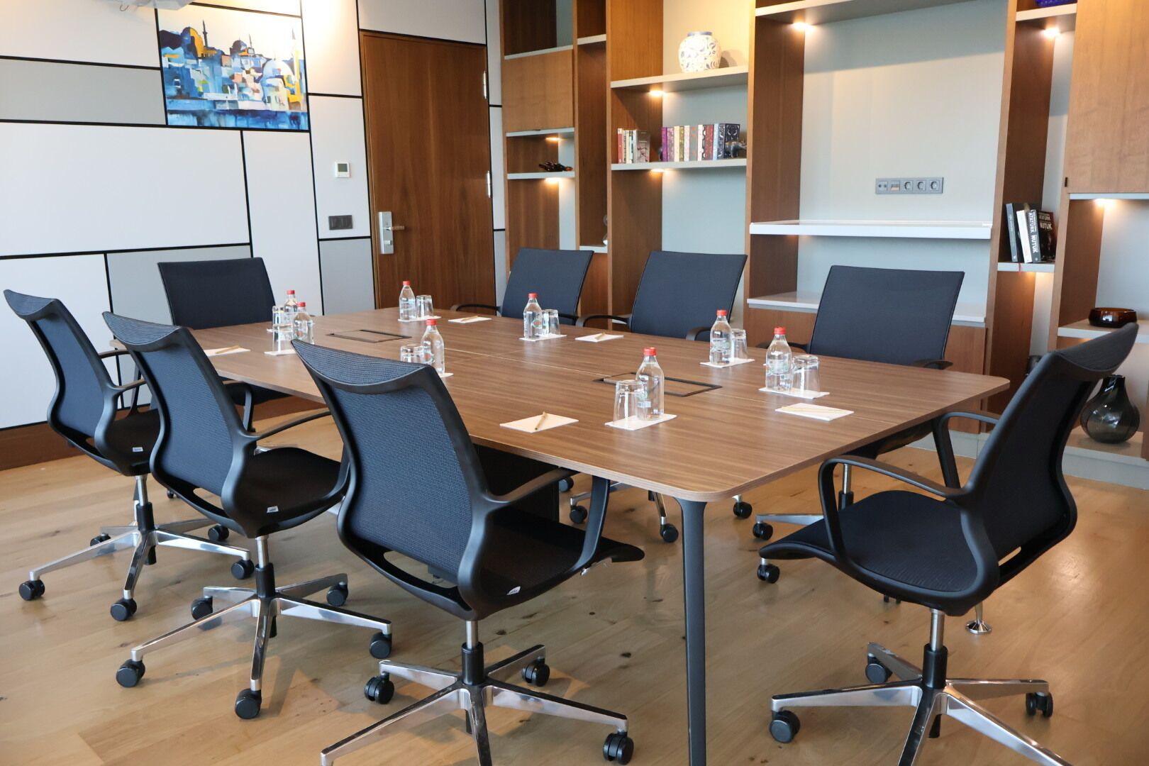 Un salón de conferencias con una mesa de madera, sillas negras y botellas de agua.