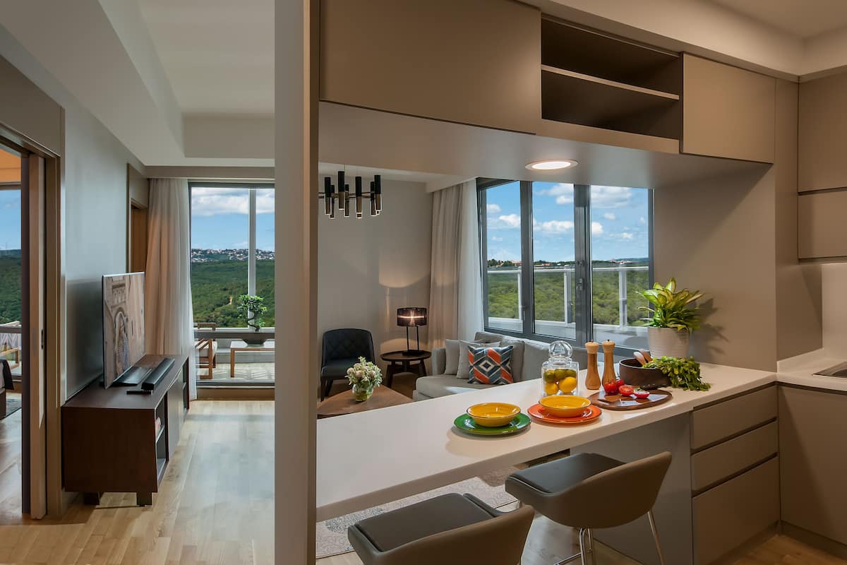 A modern kitchen with a large window, a dining area, and a view of greenery and a bridge.