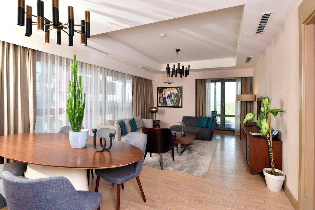 A modern hotel room with a dining area, a sofa set, a large window with curtains, and a potted plant.