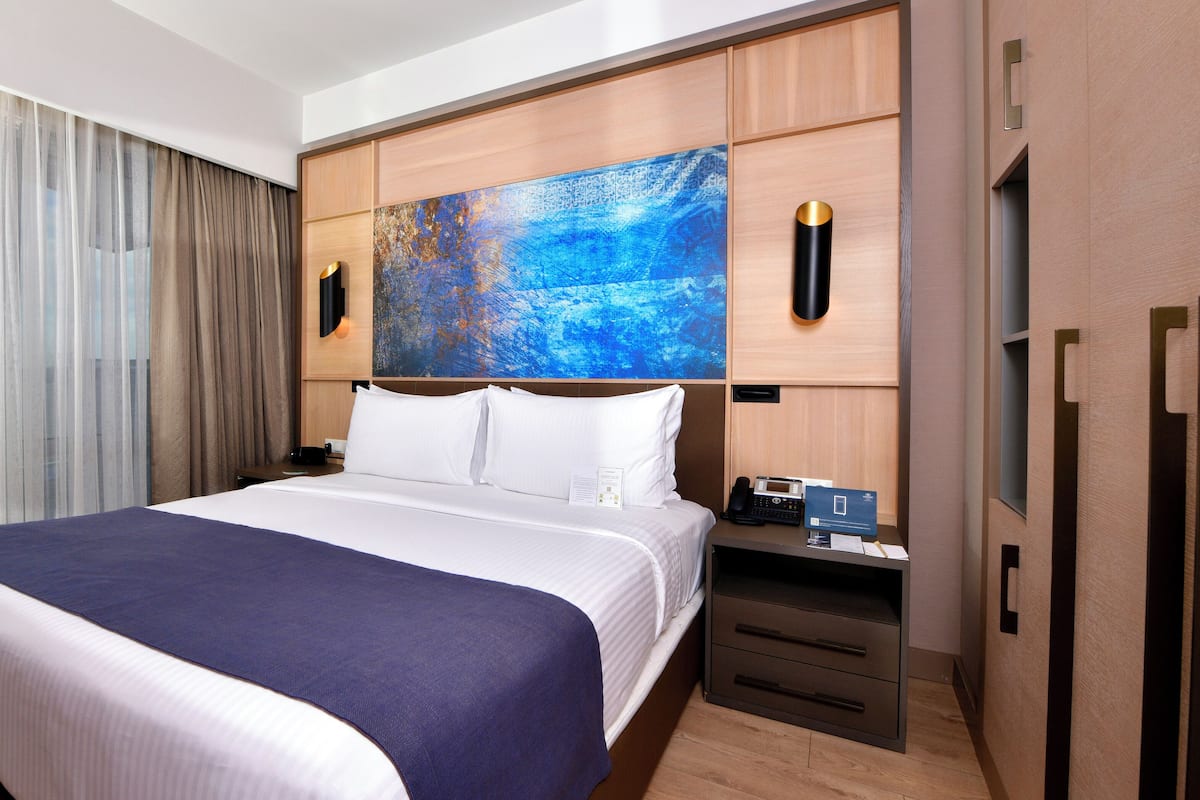A hotel room with a large bed, a nightstand with a phone and a lamp, a painting on the wall, and a view of the outdoors through the window.