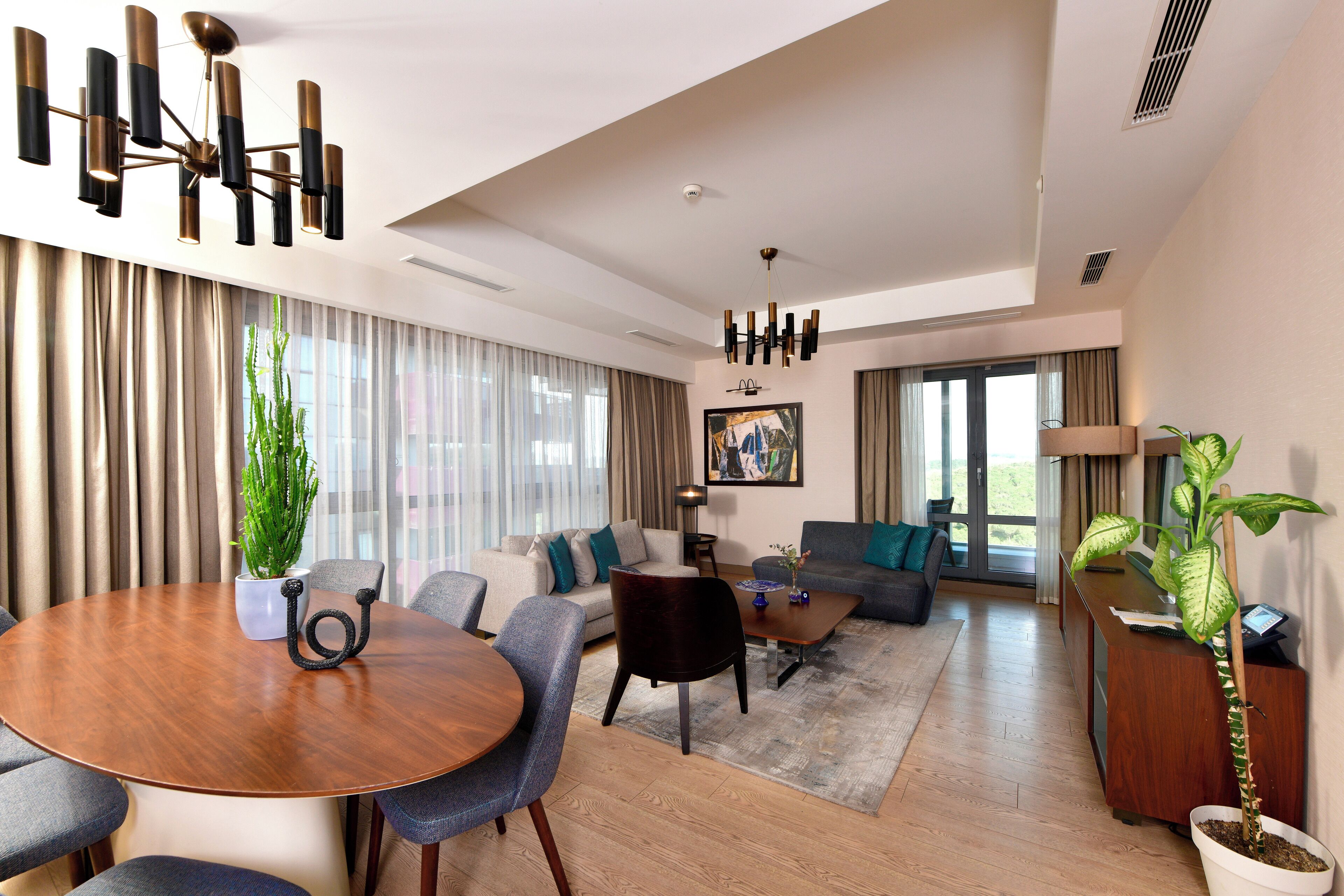 A modern living room with a round wooden table, grey sofas, and a large window with curtains.