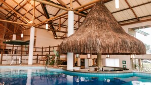 Une piscine intérieure dotée d’une structure avec toit de chaume, entourée d’un bar et d’espaces pour s’asseoir.