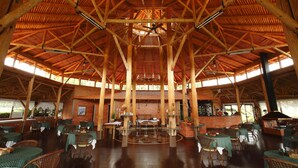 Un vaste espace intérieur doté d’un haut plafond en bois et d’un espace central ouvert. Des tables et des chaises sont disposées tout autour, et un bar est visible au fond.