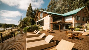 Une terrasse en bois avec des chaises longues et une table, entourée d’une maison dotée d’un toit végétalisé et de grandes fenêtres.