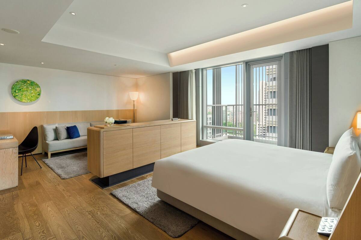 A modern hotel room with a large bed, a wooden dresser, a desk, and a view of the city through large windows.